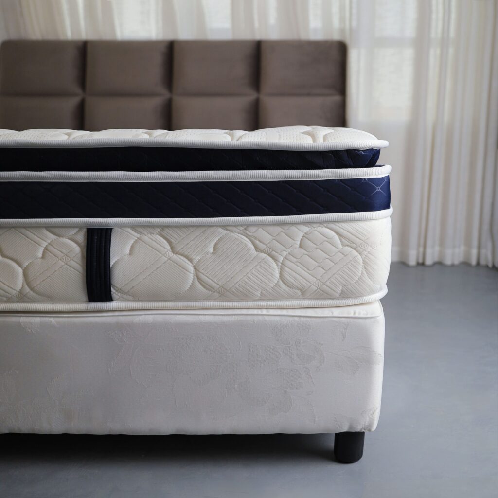 سرير أنيق مع مرتبة جديدة بيضاء وسميكة، مزود بظهر مبطن مقسم إلى مربعات، موضوع في غرفة نوم بإضاءة طبيعية وستائر بيضاء شفافة، ويظهر السرير على أرضية رمادية ناعمة.