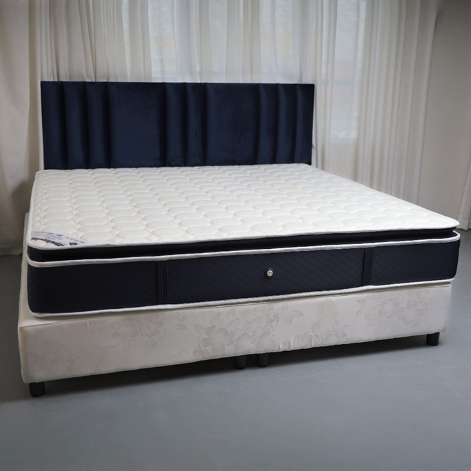 مرتبة بلون أزرق داكن بطبقة علوية بيضاء مبطنة ومنقوشة، موضوعة على قاعدة سرير بيضاء بنقش زهري خفيف، وخلفها ستائر شفافة تسمح بدخول الضوء الطبيعي.